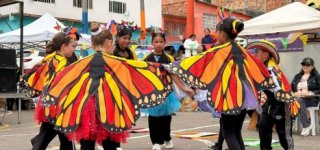 Grupo de niñas y jóvenes participa en una presentación cultural con coloridas alas de mariposa en un espacio comunitario al aire libre.